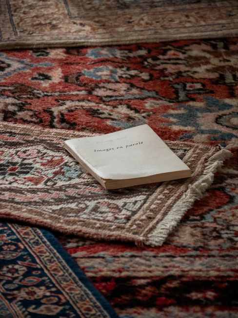 A woman with curly hair, wearing a beige jacket, dark jeans, and white shoes, is performing surface cleaning with a vacuum cleaner on a large, ornate, patterned rug in a living room. The vacuum cleane