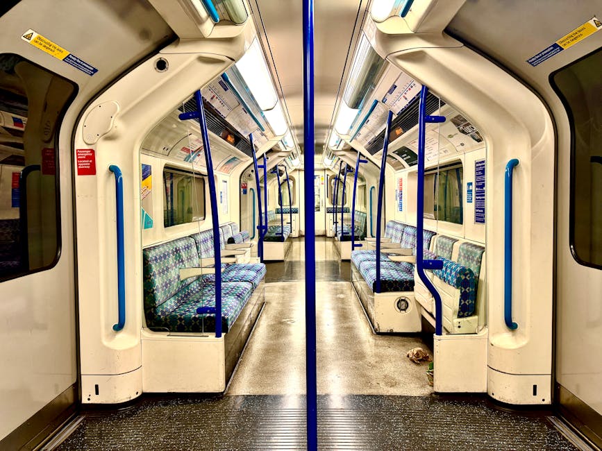 Interior view of a clean, well-maintained train carriage near Cockfosters tube station, featuring a central aisle with patterned fabric-covered seats arranged in pairs along both sides. The seats are 